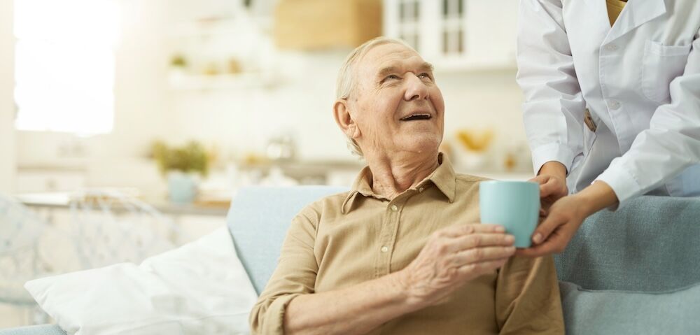 Smiling senior citizen looking at her nurse and going to drink a hot drink at home Senior man receiving care from his senior memory care community