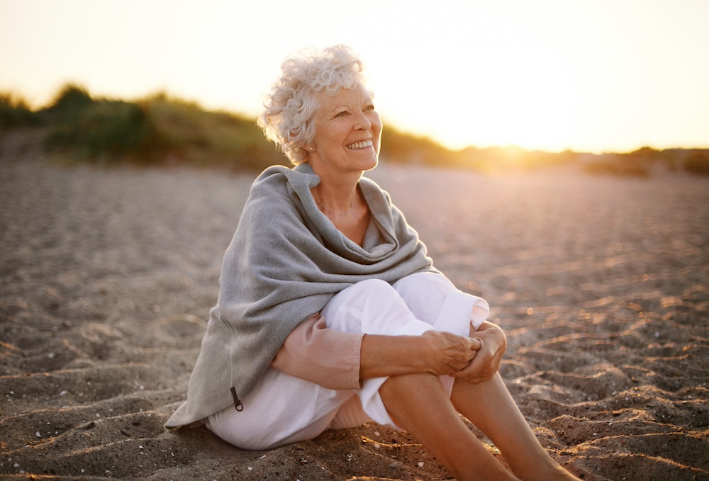 Happy retired woman wearing shawl sitting relaxed on sand at the beach